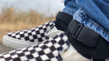 Close-up of a person wearing blue jeans, black-and-white checkered fuzzy slip-on shoes, and an ankle monitor on their right leg outdoors.