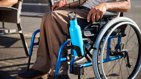 A person sitting in a blue wheelchair outdoors, holding a newspaper, with a bright blue water bottle attached to the wheelchair frame.