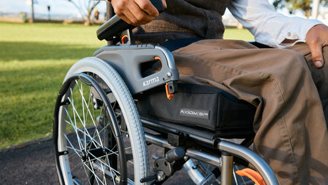 Close-up of a person’s hand resting on the armrest of a wheelchair outdoors, with grass and trees visible in the background. The wheelchair brand Karma is visible on the frame.