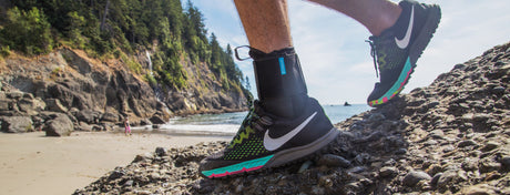 A person hiking on a rocky beach wearing black and green athletic shoes. The background shows tree-covered cliffs and the ocean.