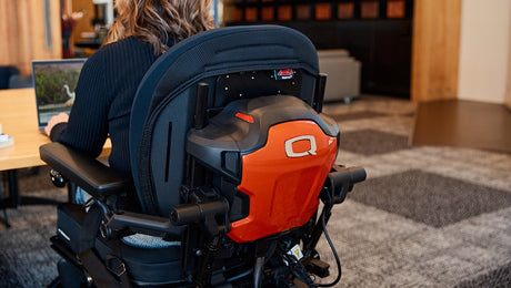 Person sitting in a motorized wheelchair featuring a sleek, orange backing with a bold Q logo. They are positioned at a wooden desk, using a laptop in a modern office environment.