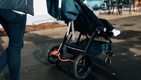 A person pushes a stroller with a young child sitting inside on a paved path outdoors. The child’s legs and white shoes are visible.