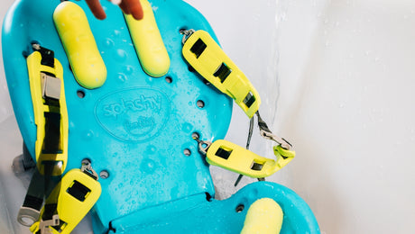 A blue adaptive bath chair with yellow straps and padding, wet from water, is set up inside a bathtub. The brand name Splashy is visible on the seat.