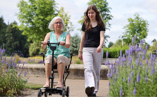 An older woman with white hair and glasses uses a motorized wheelchair on a garden path, accompanied by a young woman walking beside her in a park setting with purple flowers.